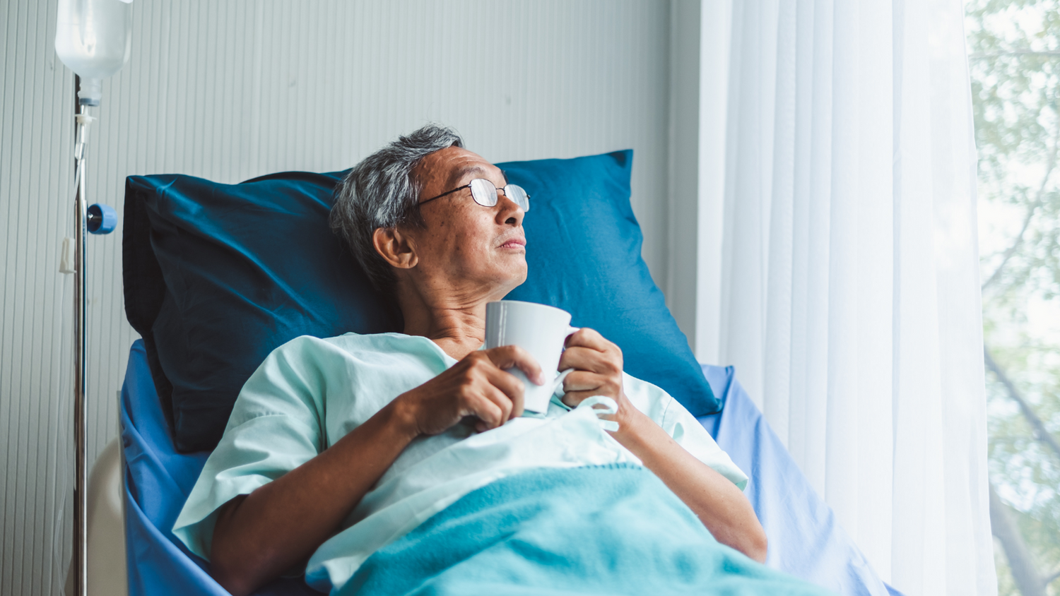 A man in a hospital bed holding a mug looking out a window